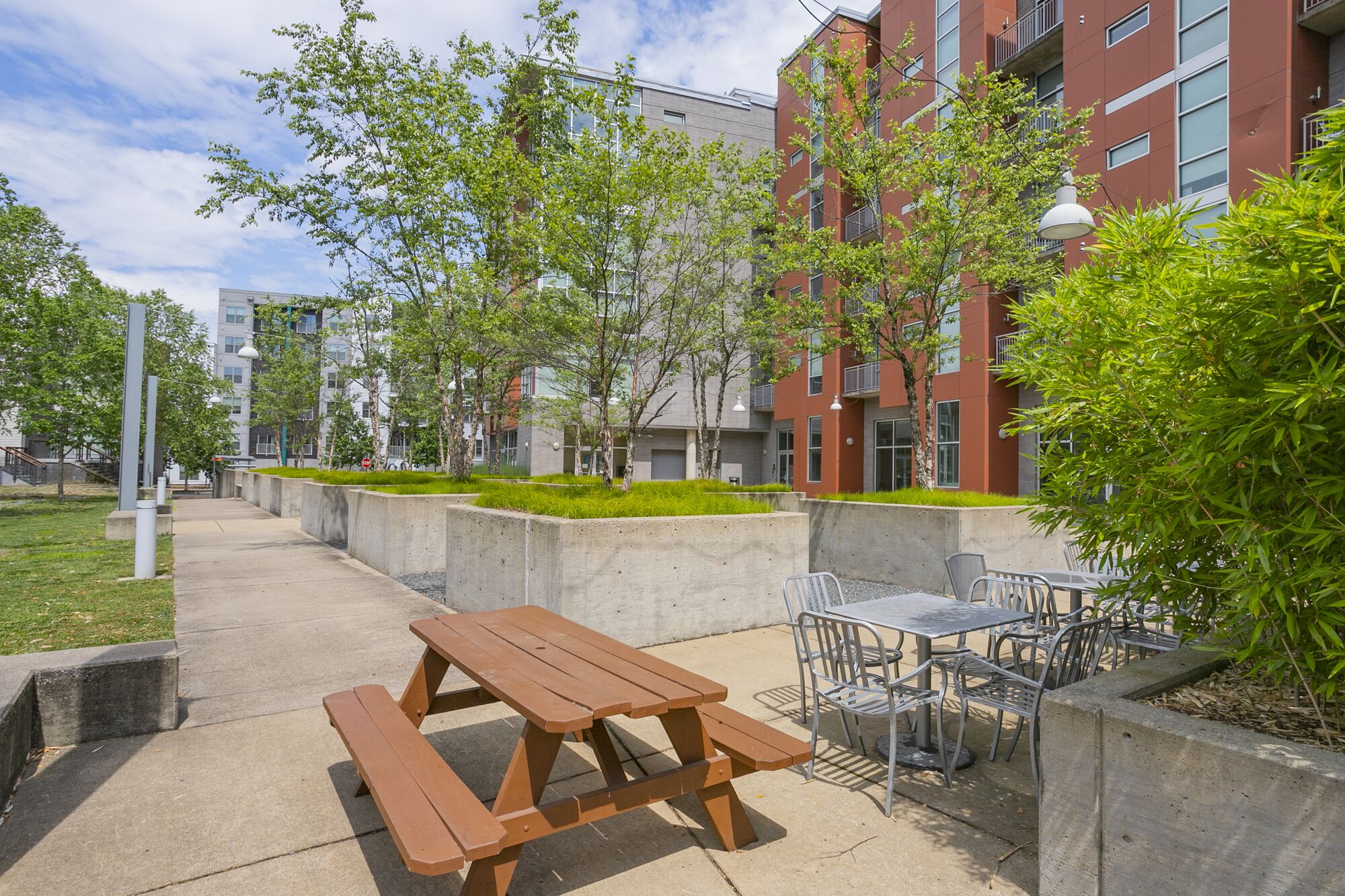 An outdoor patio with picnic tables and chairs, surrounded by greenery and modern buildings.