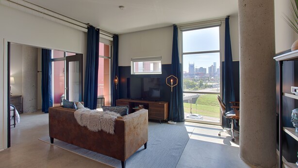 Amplia sala de estar con un ventanal grande, un sofá, un televisor y vistas del horizonte de la ciudad.