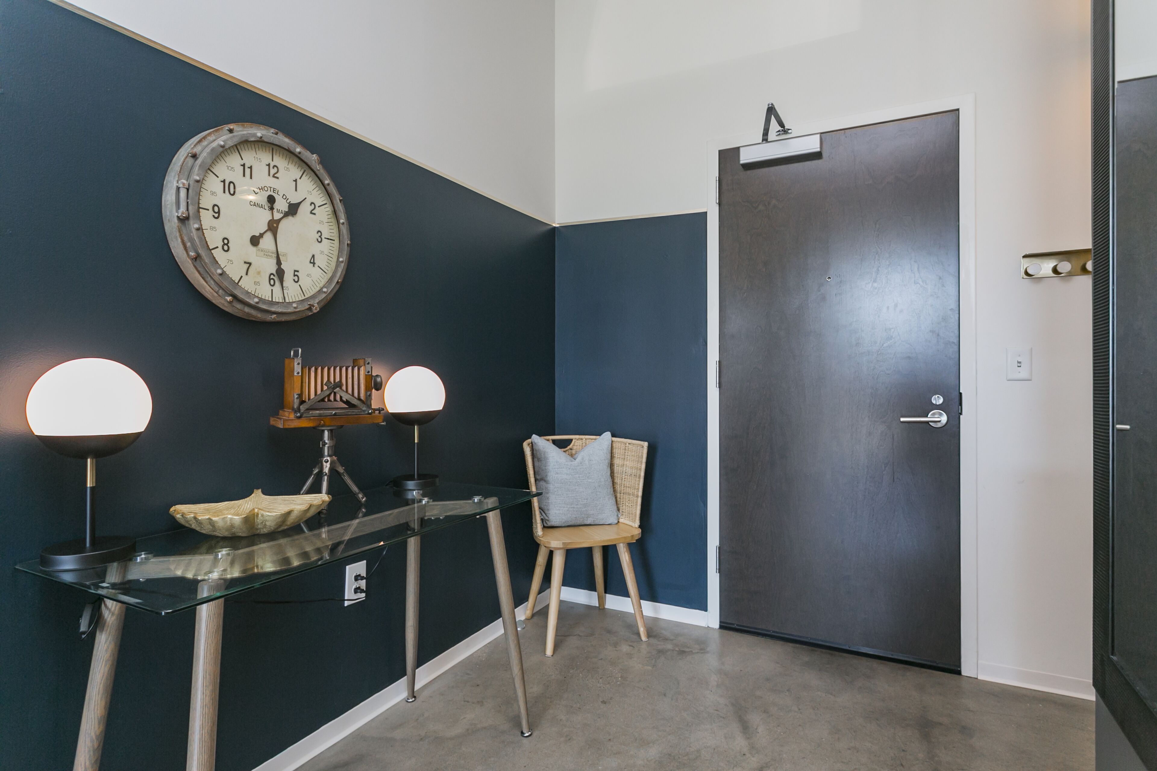 A room with a dark blue wall, a round clock, a door, a chair, and a table with two lamps.