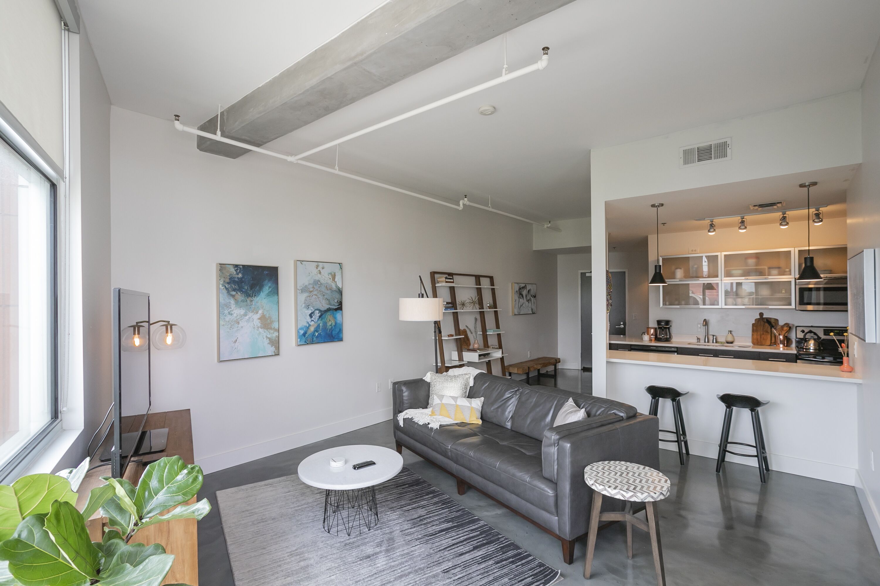 Una sala moderna con un sofá de cuero gris, una mesa de centro redonda y una zona de cocina con taburetes de bar.