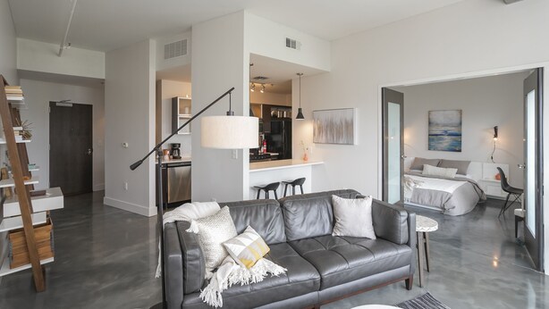 Una sala moderna con un sofá de cuero gris, un comedor con cocina al fondo y un dormitorio con cama y sillón.