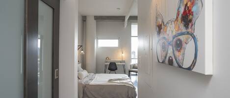 A narrow hallway leading to a bedroom with a bed, a desk, and a chair.