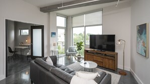 Una sala de estar moderna con un sofá gris, una mesa de centro, un televisor sobre un mueble de madera y un ventanal con vistas al exterior.