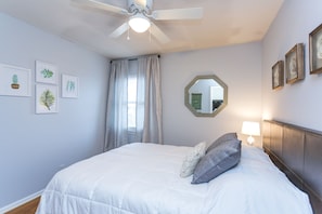 A neatly made bed with a white comforter and two pillows, a ceiling fan, and a window with curtains.