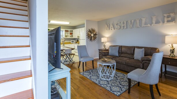 A living room with a brown leather sofa, two grey armchairs, a coffee table, and a wall-mounted TV. A decorative sunburst wall art piece and a lamp are also visible.