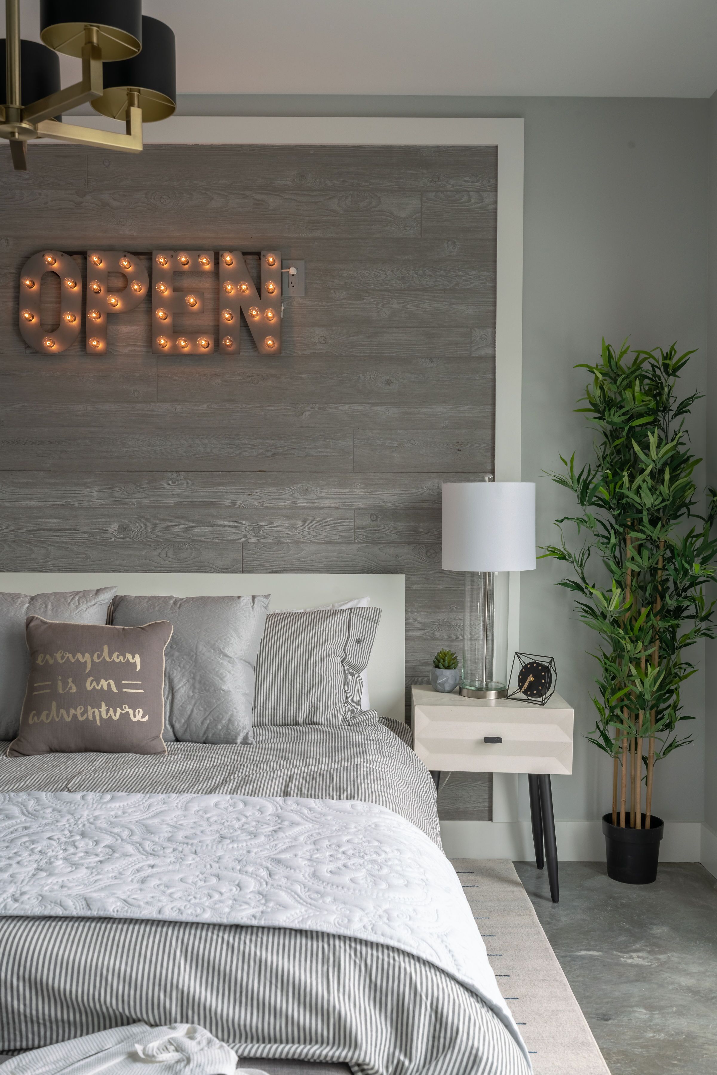 A modern bedroom with a bed, pillows, a nightstand, a lamp, a clock, and a wall-mounted sign that reads "OPEN".