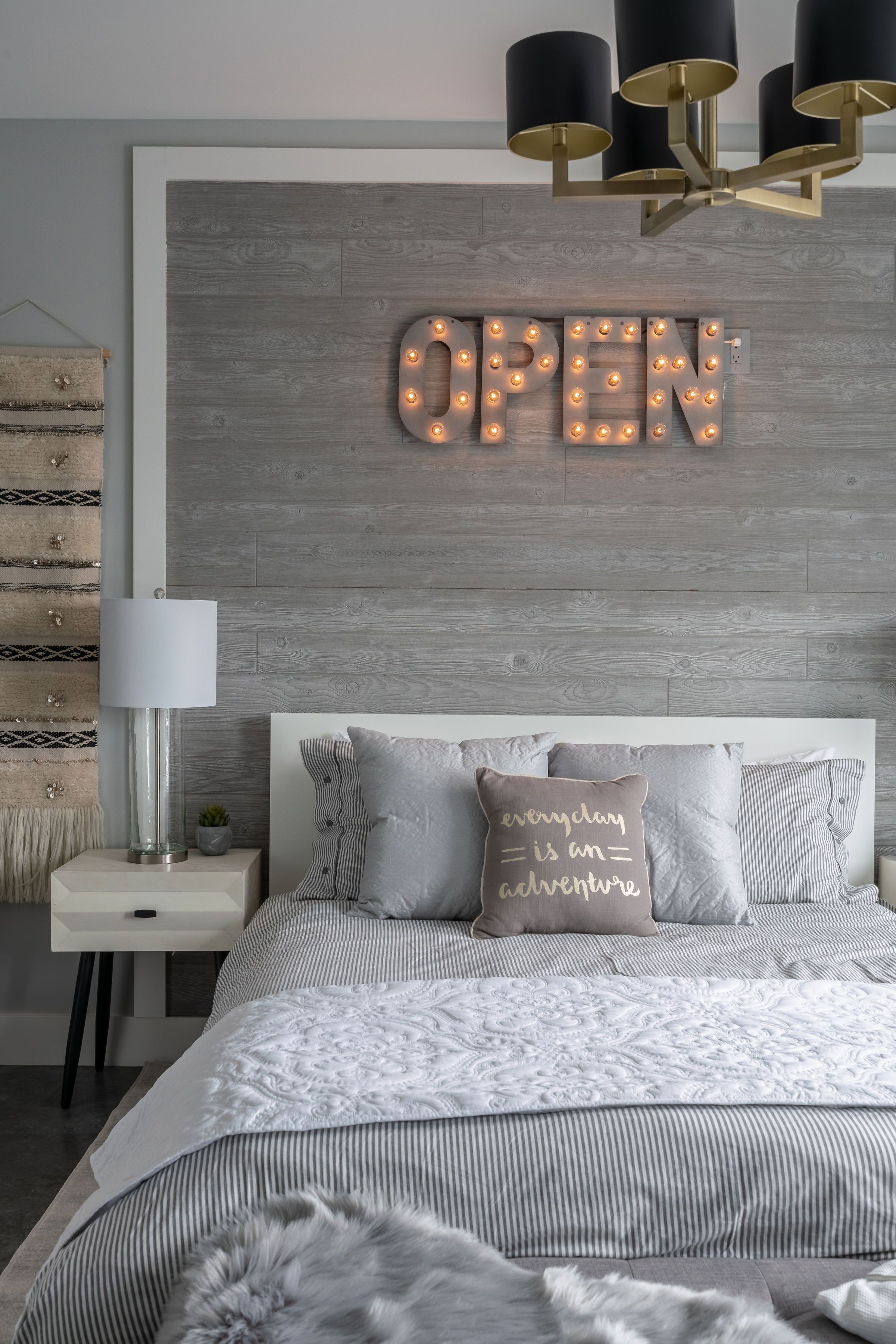 Une chambre moderne avec un lit, une table de chevet, une lampe et un panneau fixé au mur sur lequel on peut lire « OUVERT ».