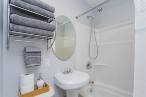 A bathroom with a white sink, a mirror, a towel rack with towels, a toilet paper holder, and a shower. - Quaint Music City Bungalow Retreat (Nashville)