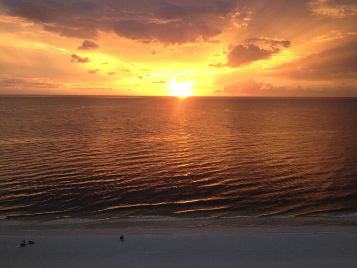A sunset over a beach with calm waves and a few people on the sand.