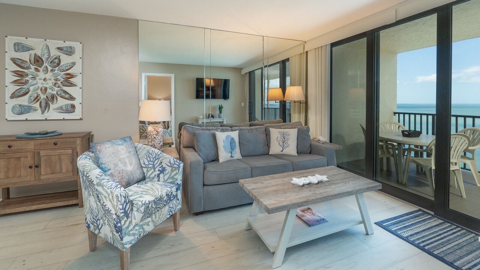 A modern living room with a sofa, armchair, and coffee table. There is a television mounted on the wall and a balcony with outdoor seating.
