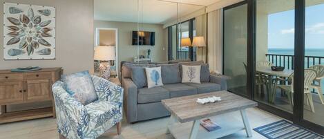 A modern living room with a sofa, armchair, and coffee table. There is a television mounted on the wall and a balcony with outdoor seating.