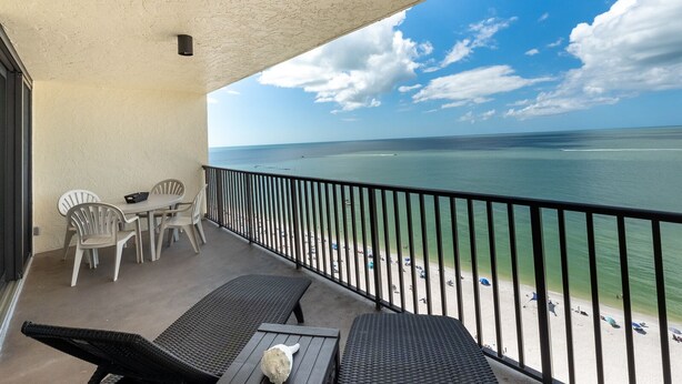 A balcony with a view of the beach, a table with chairs, and a lounge chair.