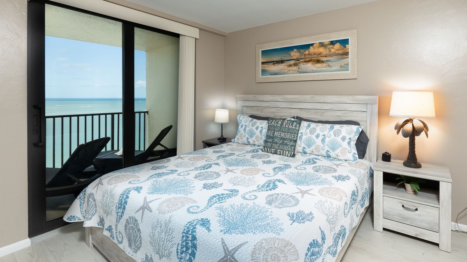 A bedroom with a bed, a nightstand, a lamp, a chair, and a view of the ocean.