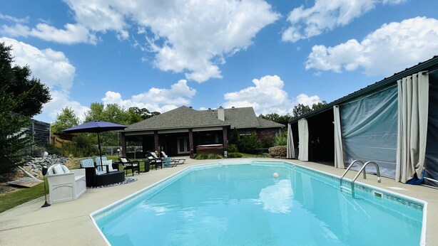 Ein rechteckiger Swimmingpool mit klar blauem Wasser, umgeben von einer Betonplattform. Liegestühle und ein überdachter Patio-Bereich mit einer Pergola sind im Hintergrund zu sehen.