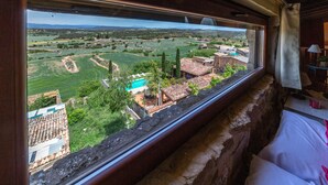 A room with a view of a rural landscape, stone walls, a bed, and a small table with a lamp.