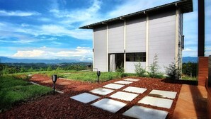 A modern house with a stone pathway leading to the entrance, surrounded by lush greenery and a mountainous backdrop.