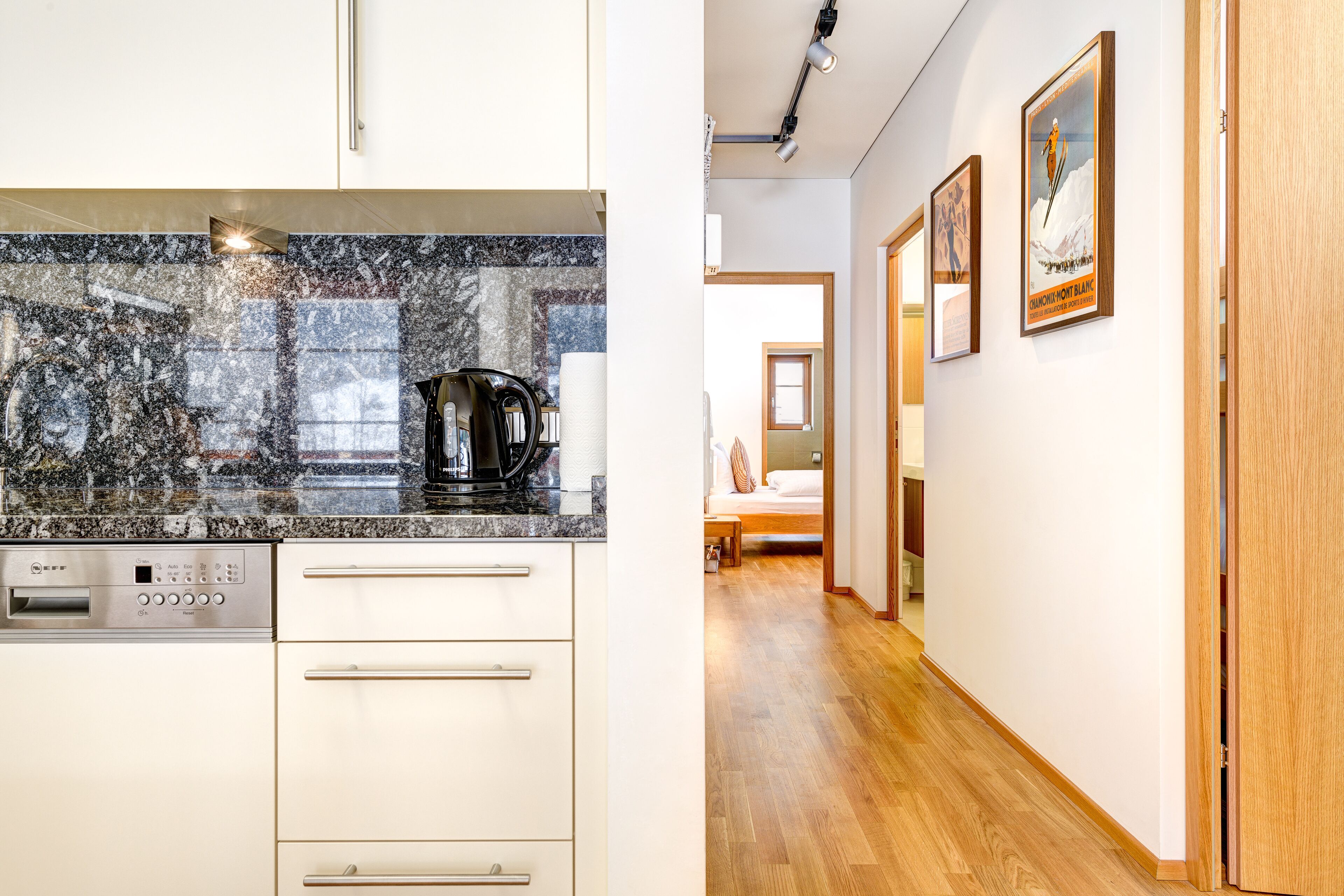 A modern kitchen with a granite countertop, stainless steel appliances, and a wooden floor leading to a bedroom with a bed and a nightstand.