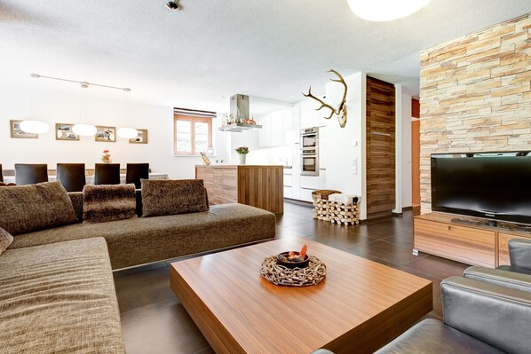 A modern living room with a sectional sofa, a wooden coffee table, a flat-screen TV, and a stone accent wall. - Mountain Lodge by A-Appartements (Brand)