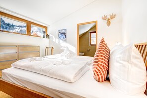 A bedroom with a wooden bed, white bedding, a striped pillow, a wooden headboard, a window with a view of a snowy landscape, and a wall-mounted mirror. - Mountain Lodge by A-Appartements (Brand)