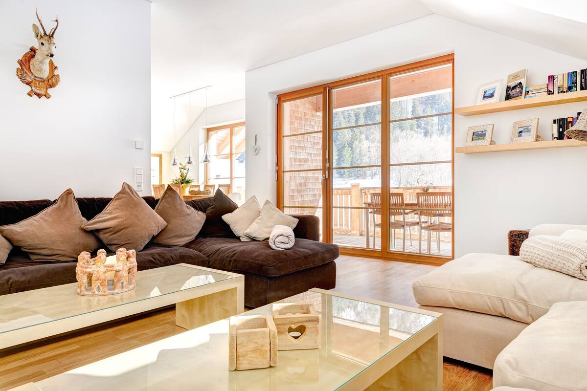 Una sala de estar moderna con un sofá marrón, una mesa de centro de vidrio y un sofá color crema. Hay estantes de madera con libros e imágenes, y un ventanal con vistas a muebles de exterior.
