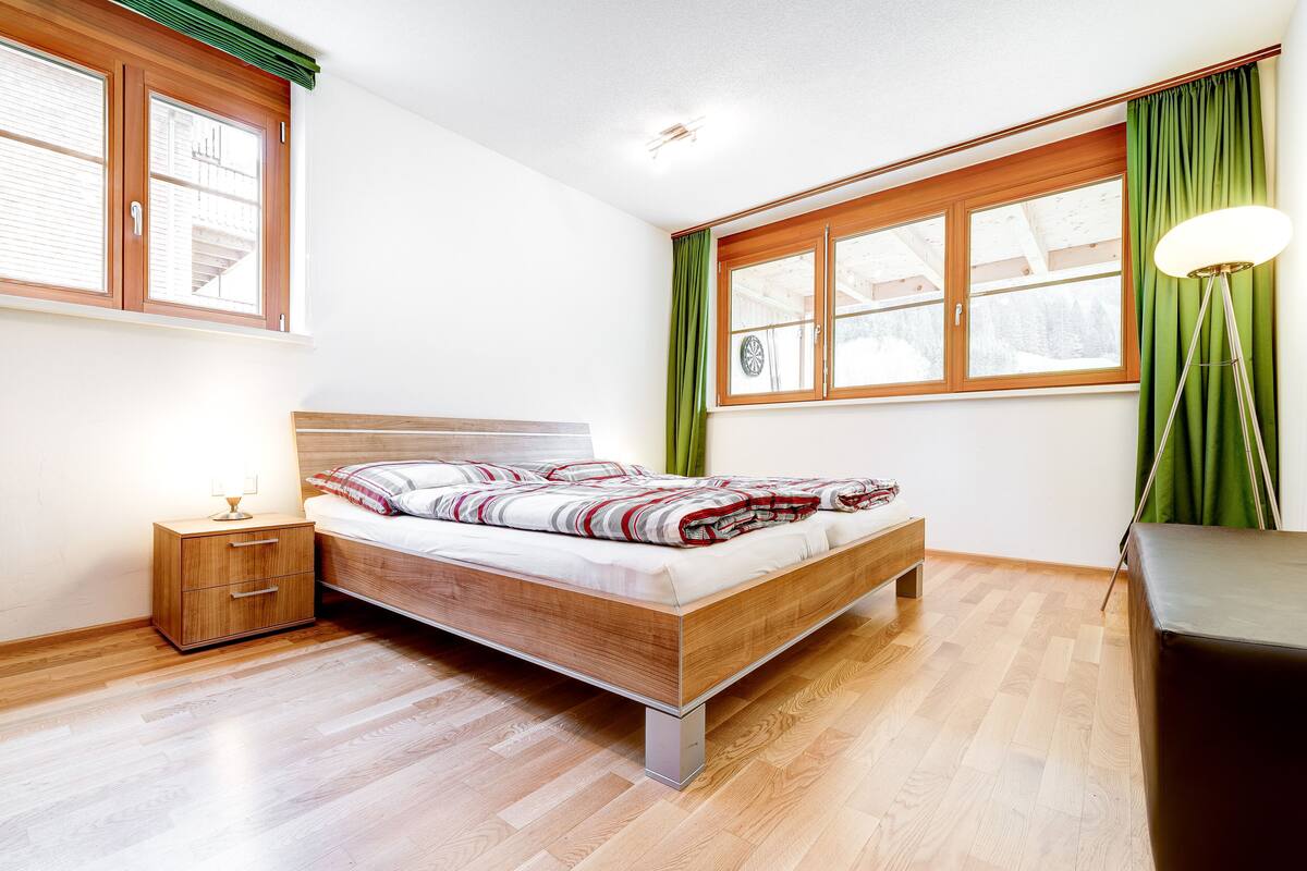 Un dormitorio moderno con una cama de madera, una mesita de noche, una lámpara de pie y ventanas amplias con cortinas.
