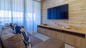 A modern living room with a large flat-screen TV mounted on a wooden wall, a sofa with patterned cushions, and a wooden entertainment unit with various items. - Apartamento beira mar - OKA Beach Resort (Ipojuca)