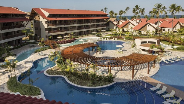 A resort with a large pool, multiple buildings with red roofs, and palm trees. - Apartamento beira mar - OKA Beach Resort (Ipojuca)