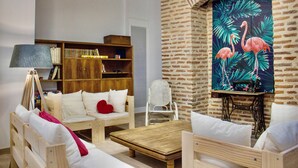 Una sala de estar acogedora con pared de ladrillo, una estantería, una mesa de centro de madera y un sofá blanco con un cojín rojo con forma de corazón.