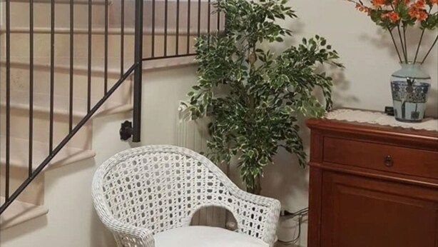 Una scala con ringhiera nera, una sedia in vimini bianco con cuscino, una pianta in vaso e un armadietto di legno.
