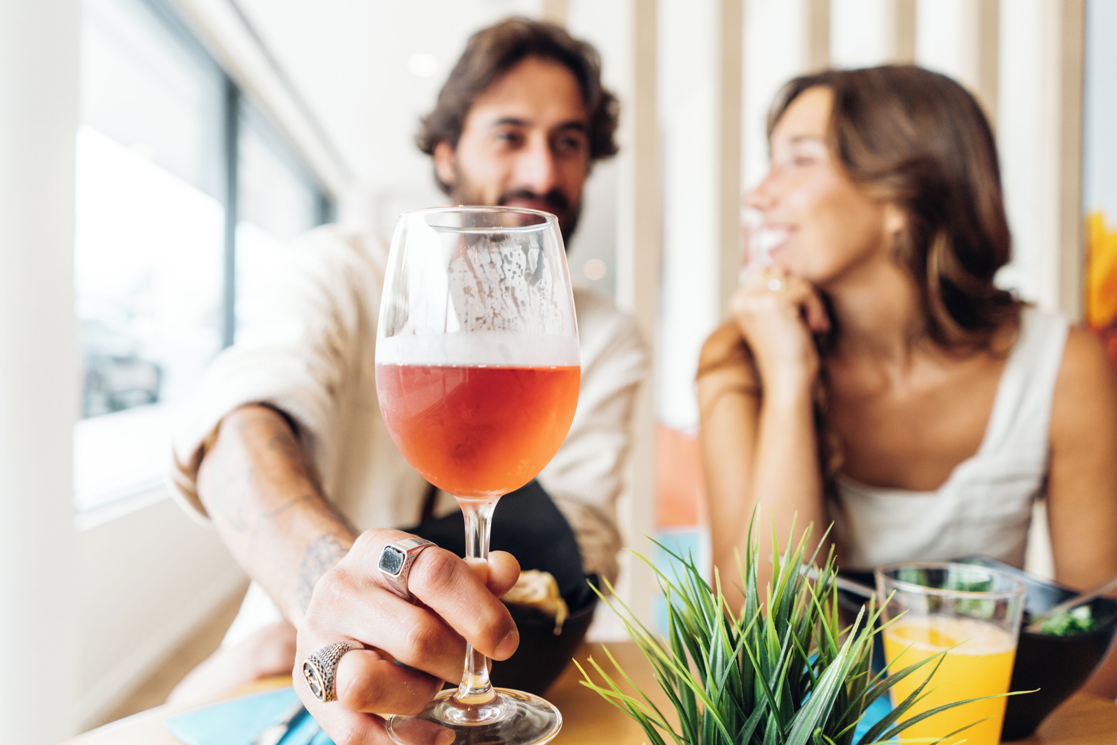 Una persona sosteniendo un vaso de bebida roja sobre una mesa con una planta y un vaso de jugo de naranja.