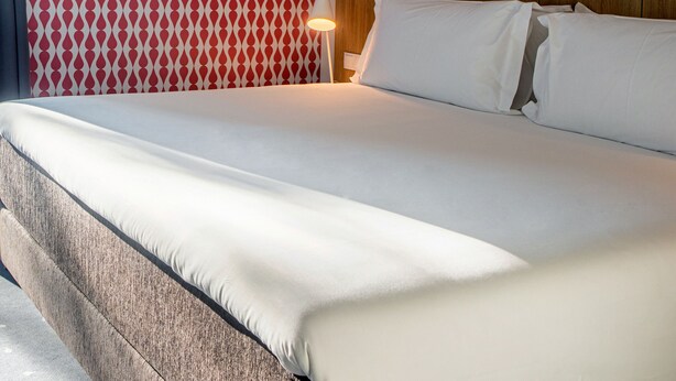 Una cama bien hecha con sábanas blancas, un cabecero de madera y una cortina estampada.