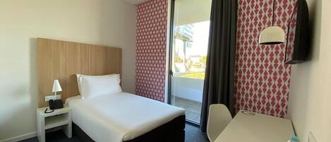Quarto de hotel com cama, escrivaninha, cadeira, televisão e janela com cortinas.