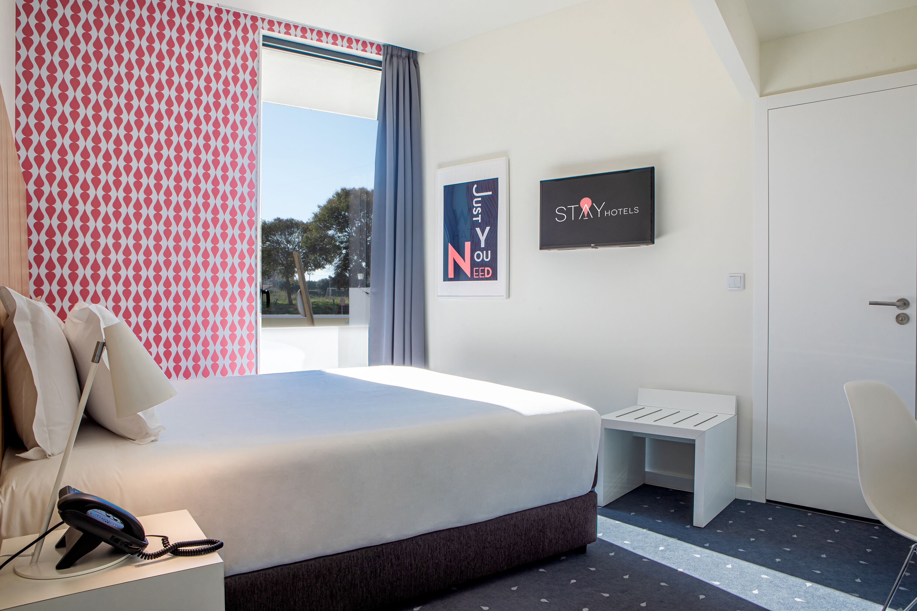 Una habitación de hotel moderna con una cama grande, una mesita de noche con lámpara, un pequeño banco blanco y un televisor en la pared.