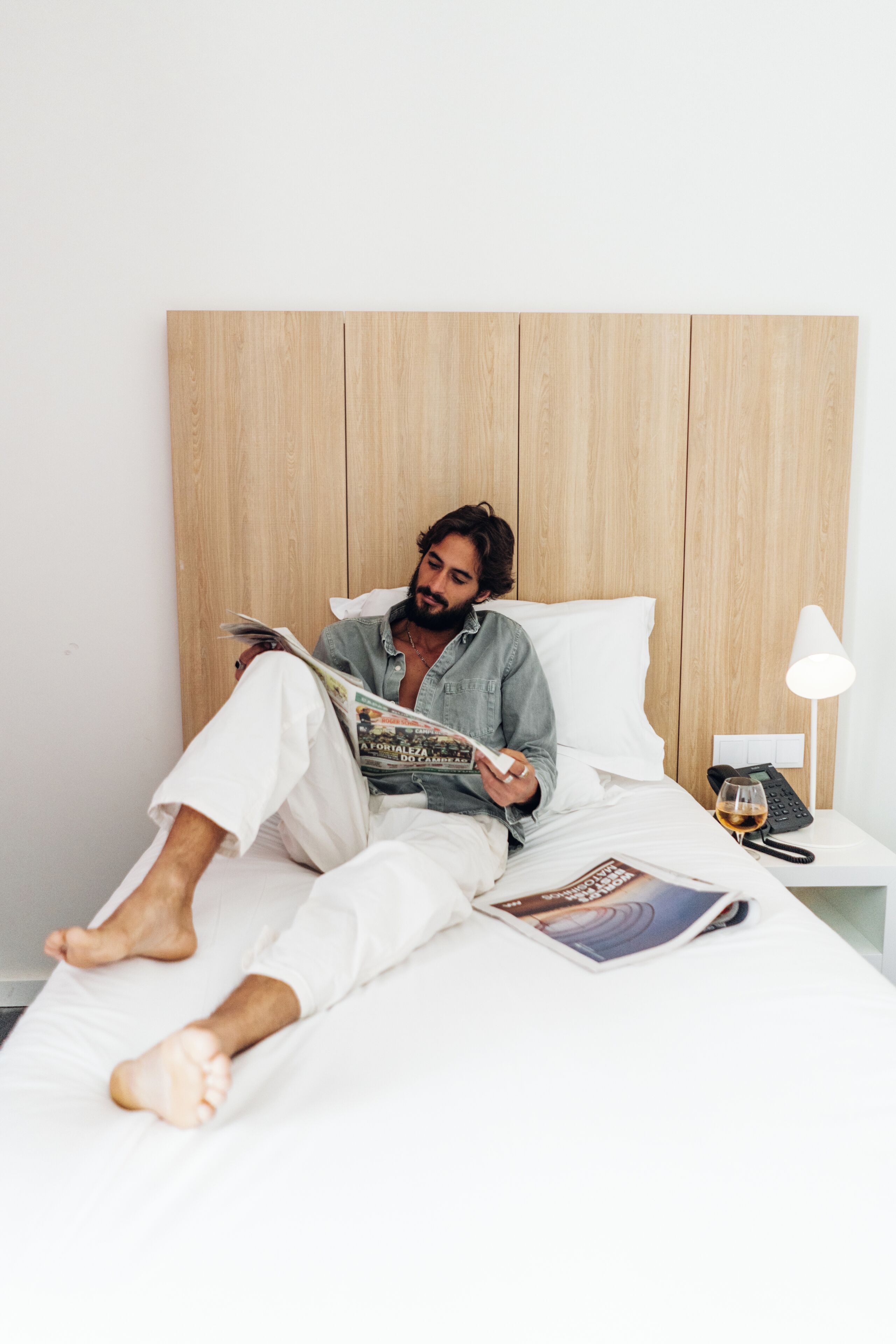 Una persona acostada en la cama leyendo una revista, con cabecera de madera, ropa de cama blanca y una lámpara de noche.