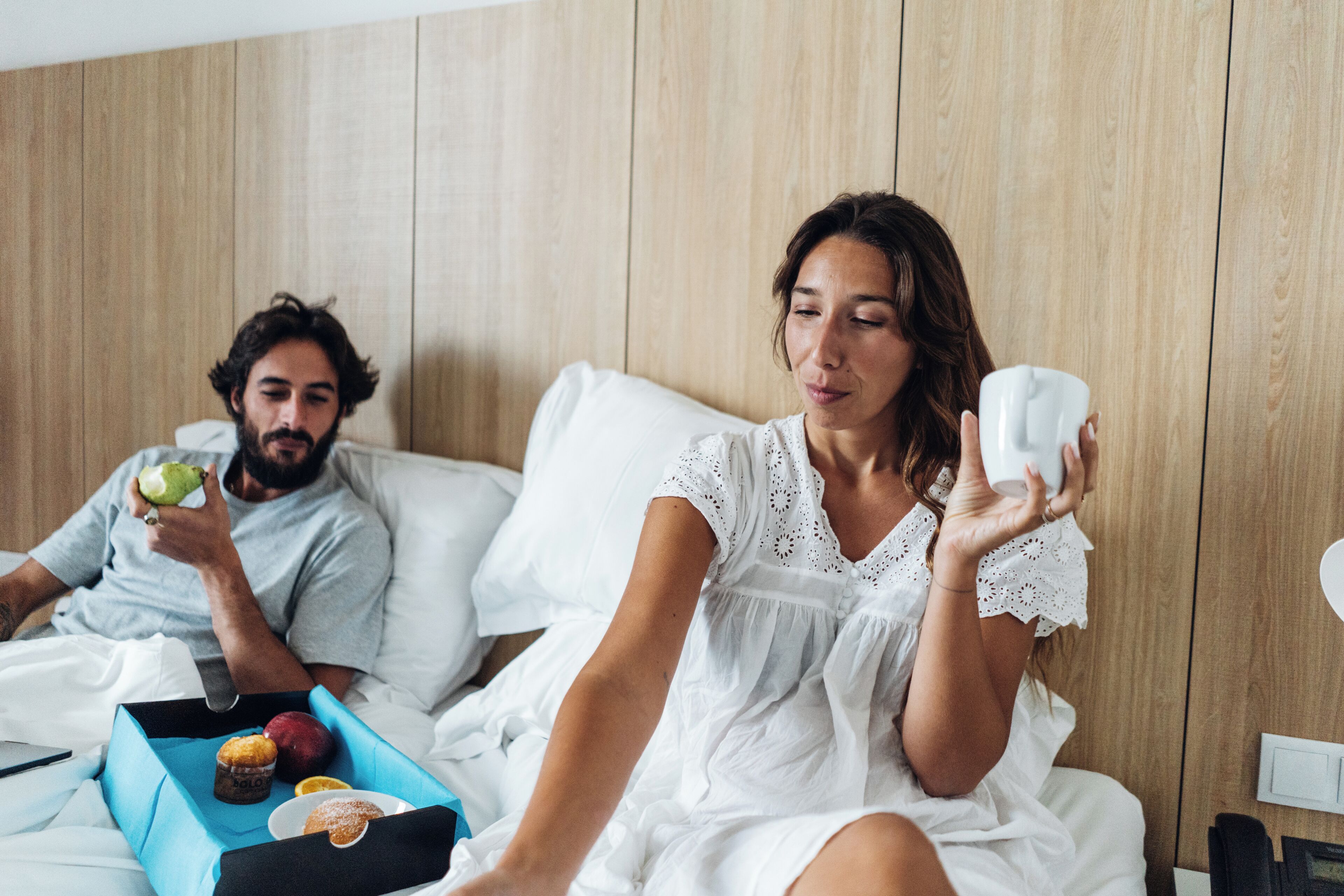 A couple in a bedroom with a wooden wall, a bedside table, and a blue tray with food items.