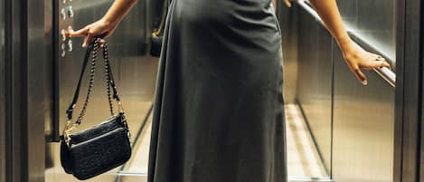 Una mujer con vestido de pie en un ascensor con paredes metálicas y pasamanos.