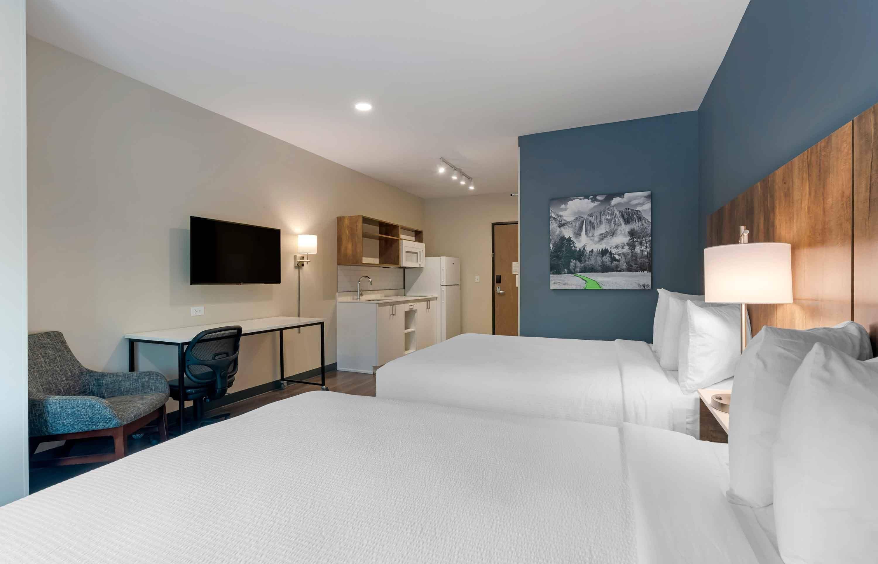 Quarto de hotel com duas camas, uma escrivaninha, uma cadeira, televisão, micro-ondas e um quadro na parede.