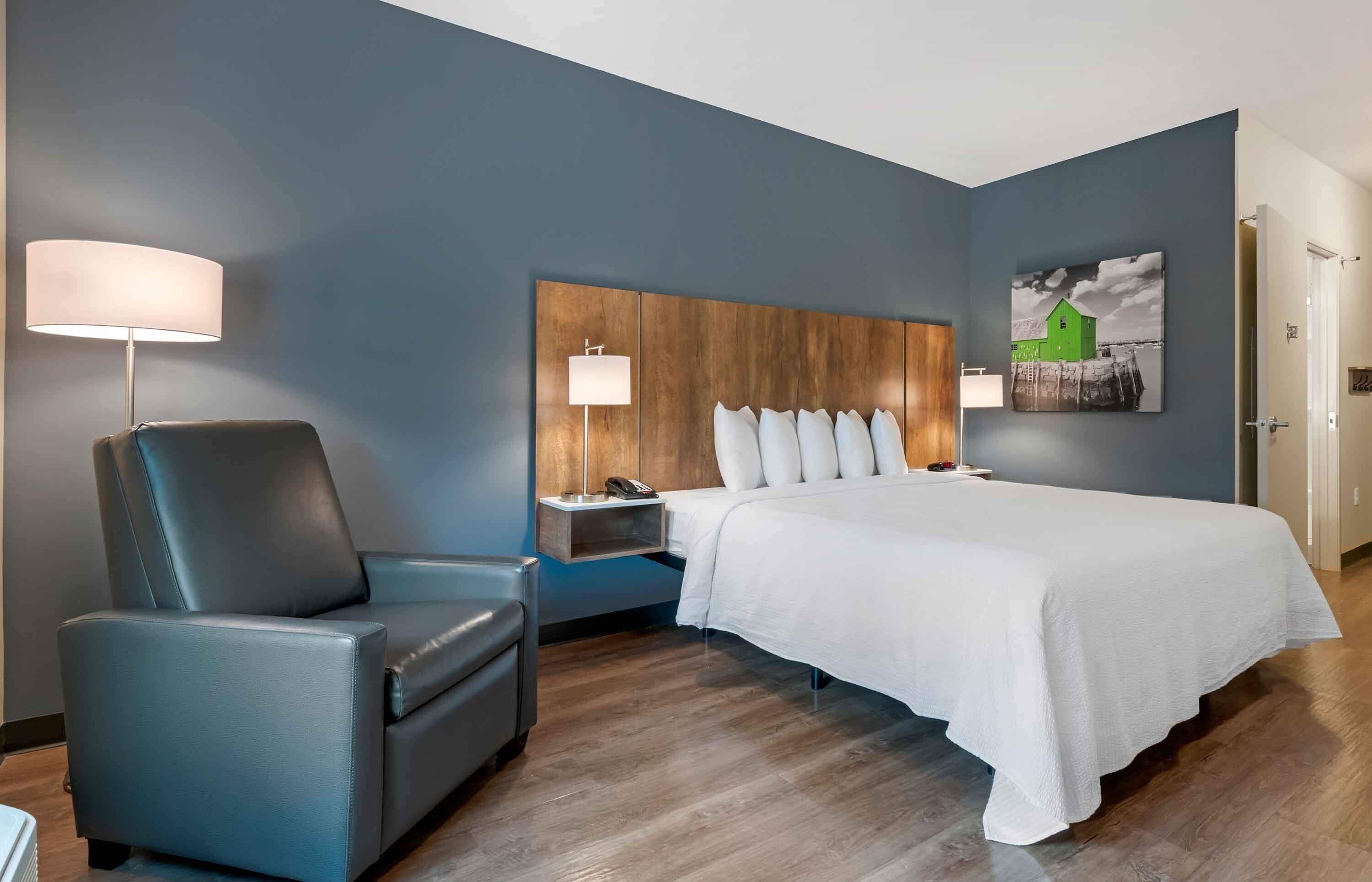 Ein Hotelzimmer mit einem Bett, einem Sessel, einem Nachttisch, einer Lampe und einem Wandbild.