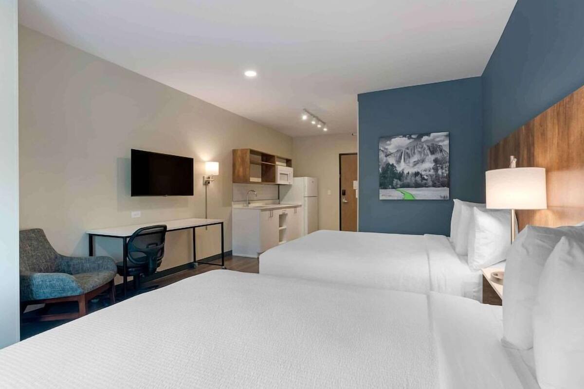 A hotel room with two beds, a desk with a chair, a flat-screen TV, a microwave, a refrigerator, and a painting on the wall.