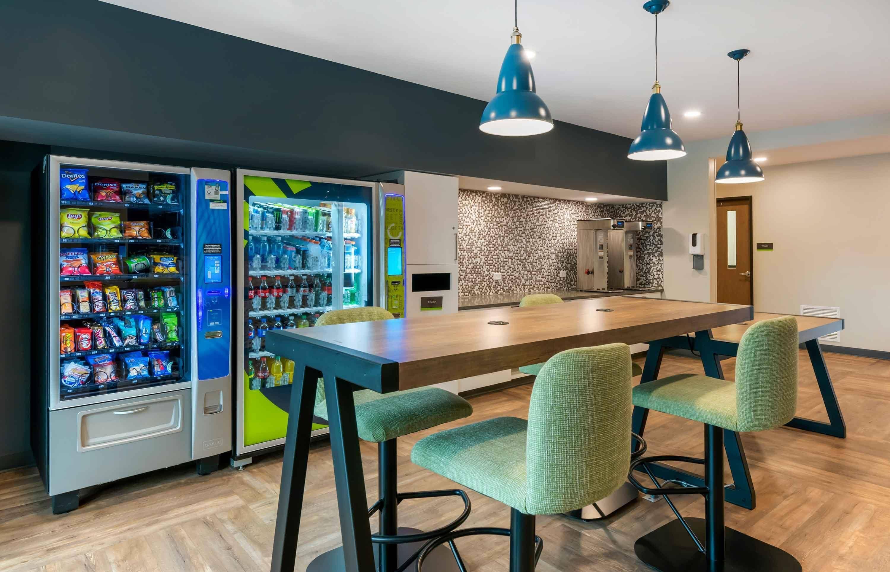 A modern hotel room with a dining area, vending machines, and a kitchen.