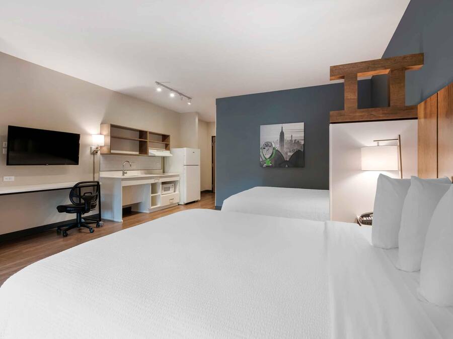 Quarto de hotel com uma cama grande, uma escrivaninha com cadeira, televisão de tela plana, um quadro na parede e um objeto decorativo de madeira.