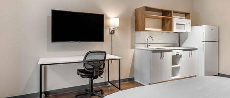 A hotel room with a bed, a desk with a chair, a flat-screen TV, a kitchenette with a microwave and refrigerator, and a wall-mounted light.