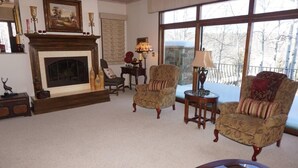 A cozy living room with a fireplace, two armchairs, a side table with a lamp, and a large window with a view of trees. - 35 Alpine Ln (Snowmass Village)
