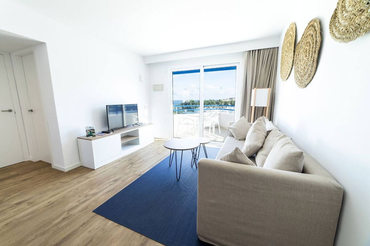 A modern living room with a sofa, a TV, a small table, and a view of the sea through large windows.
