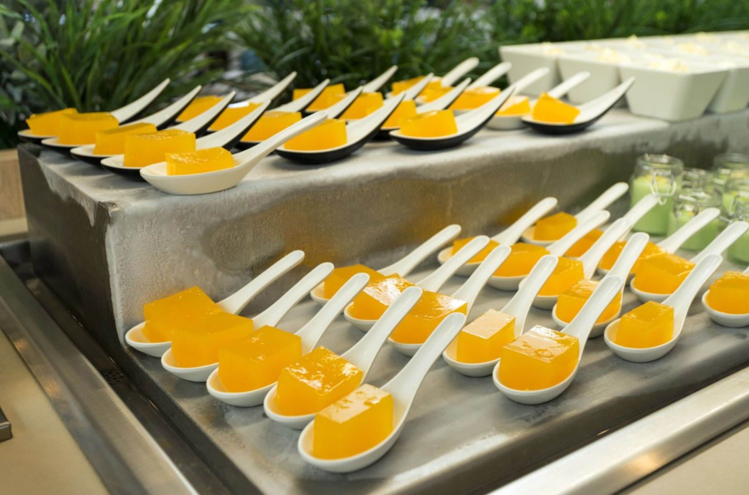 A buffet setup with multiple white spoons arranged on a metallic tray, each containing a yellow dessert.
