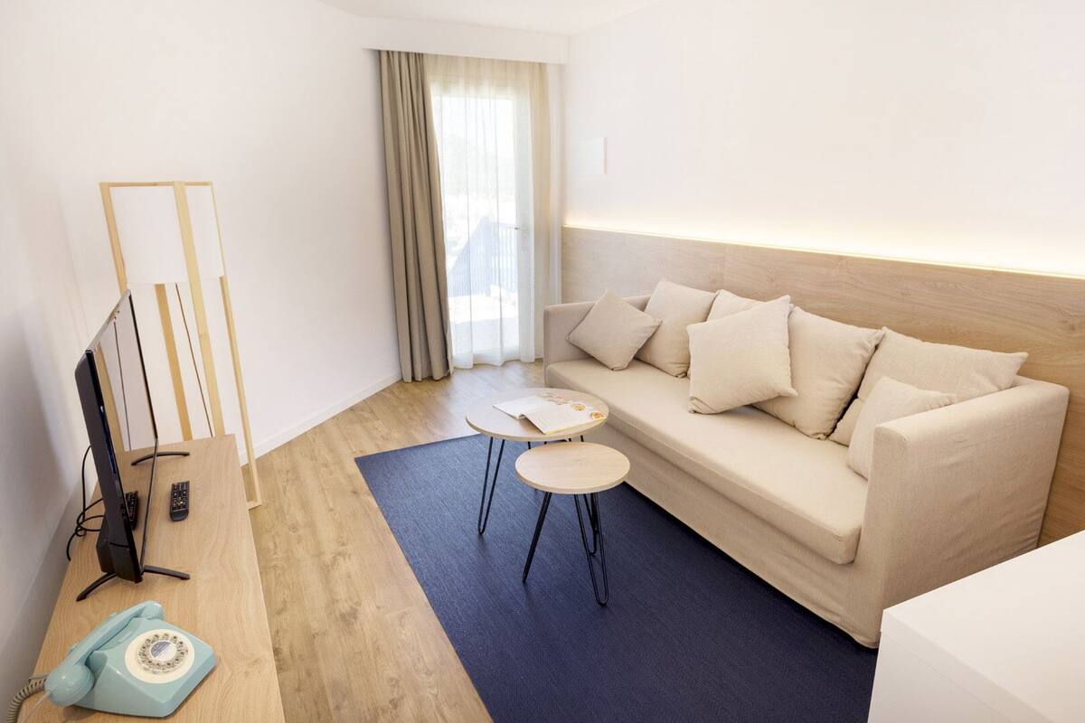 A modern hotel room with a beige sofa, two small round tables, a wooden floor, a TV, and a telephone.