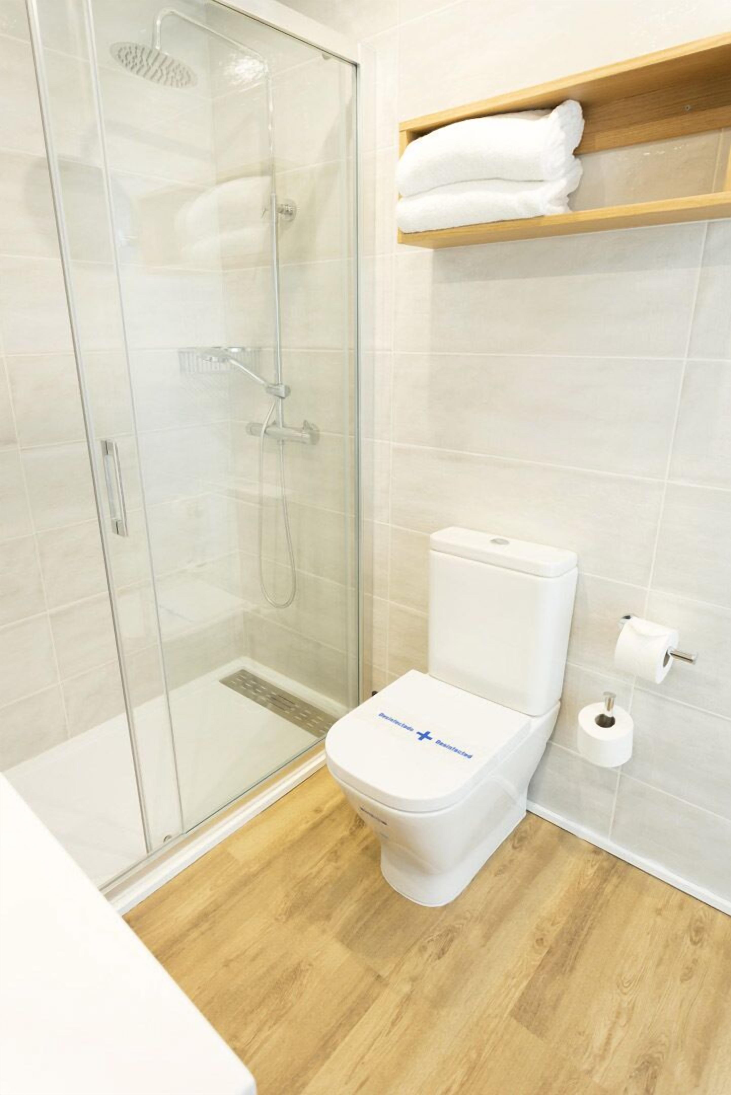 A modern bathroom with a glass-enclosed shower, a white toilet, and a wooden shelf above the toilet.