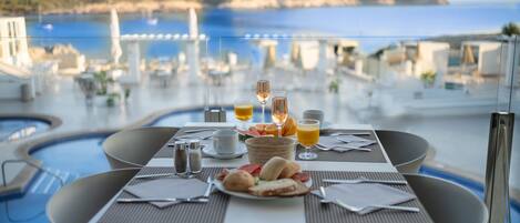 A dining table with breakfast items and drinks, overlooking a pool and a scenic coastal view.