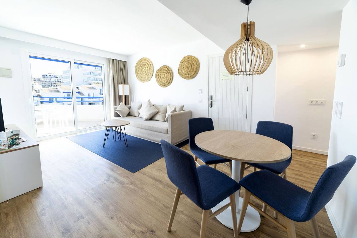 A modern living room with a sofa, a round dining table with chairs, a coffee table, and a large window with a view of buildings.
