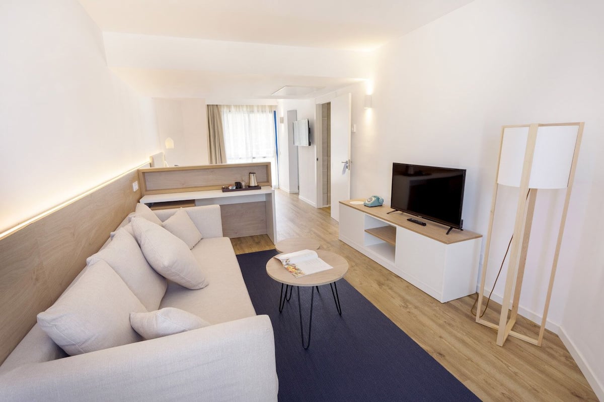 A modern hotel room with a sofa, a small table, a TV, and a bedside table.